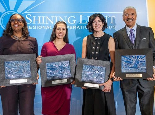 four people standing in front of a shining light regional cooperation awards backdrop holding embossed plaque awards and smiling