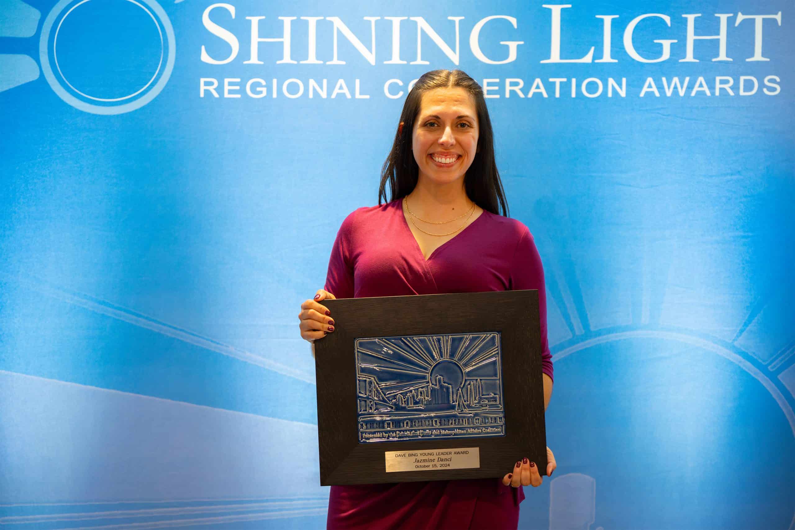 Jazzmine Dancel smiles while holding her Shining Light Regional Cooperation Award plaque in front of a blue awards backdrop.