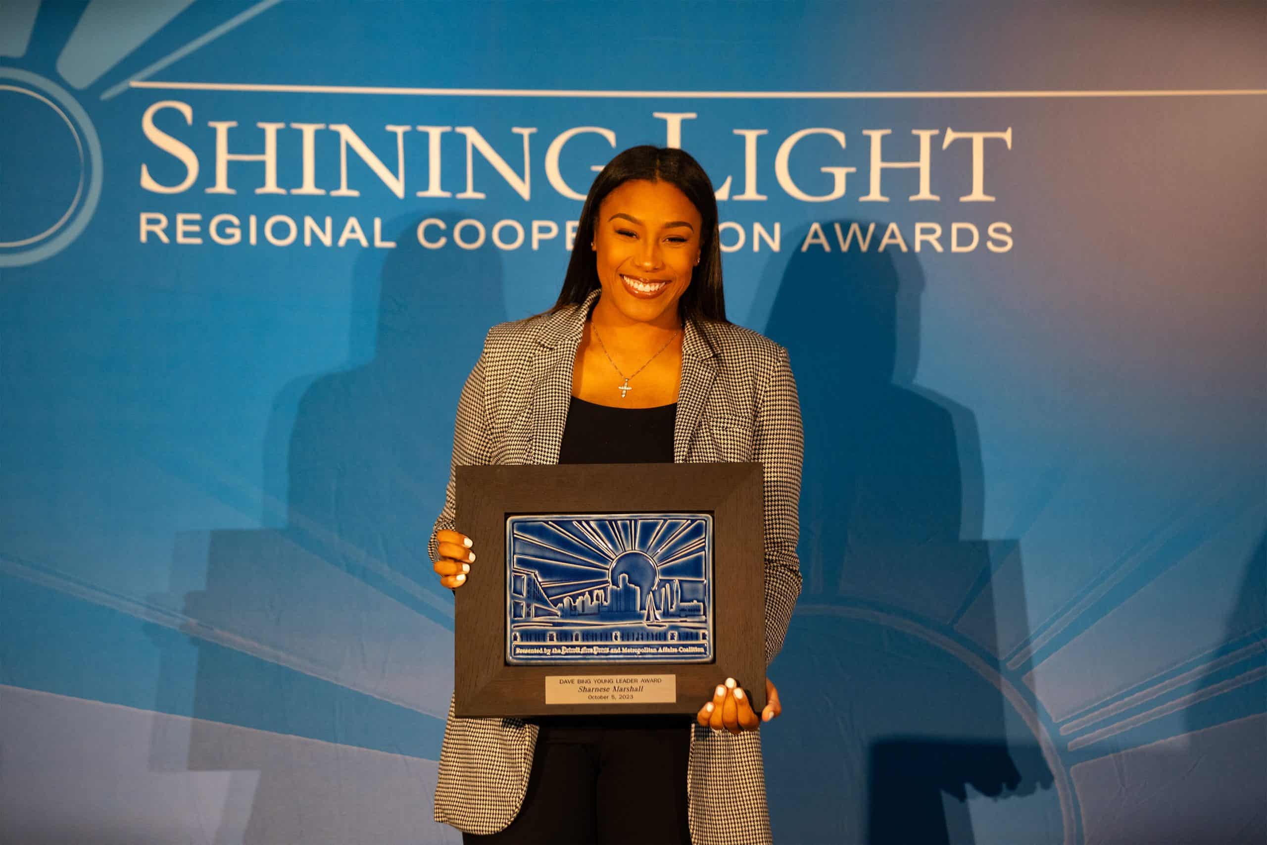 Shanese Marshall smiles while holding her Shining Light Regional Cooperation Award plaque in front of a blue awards backdrop.