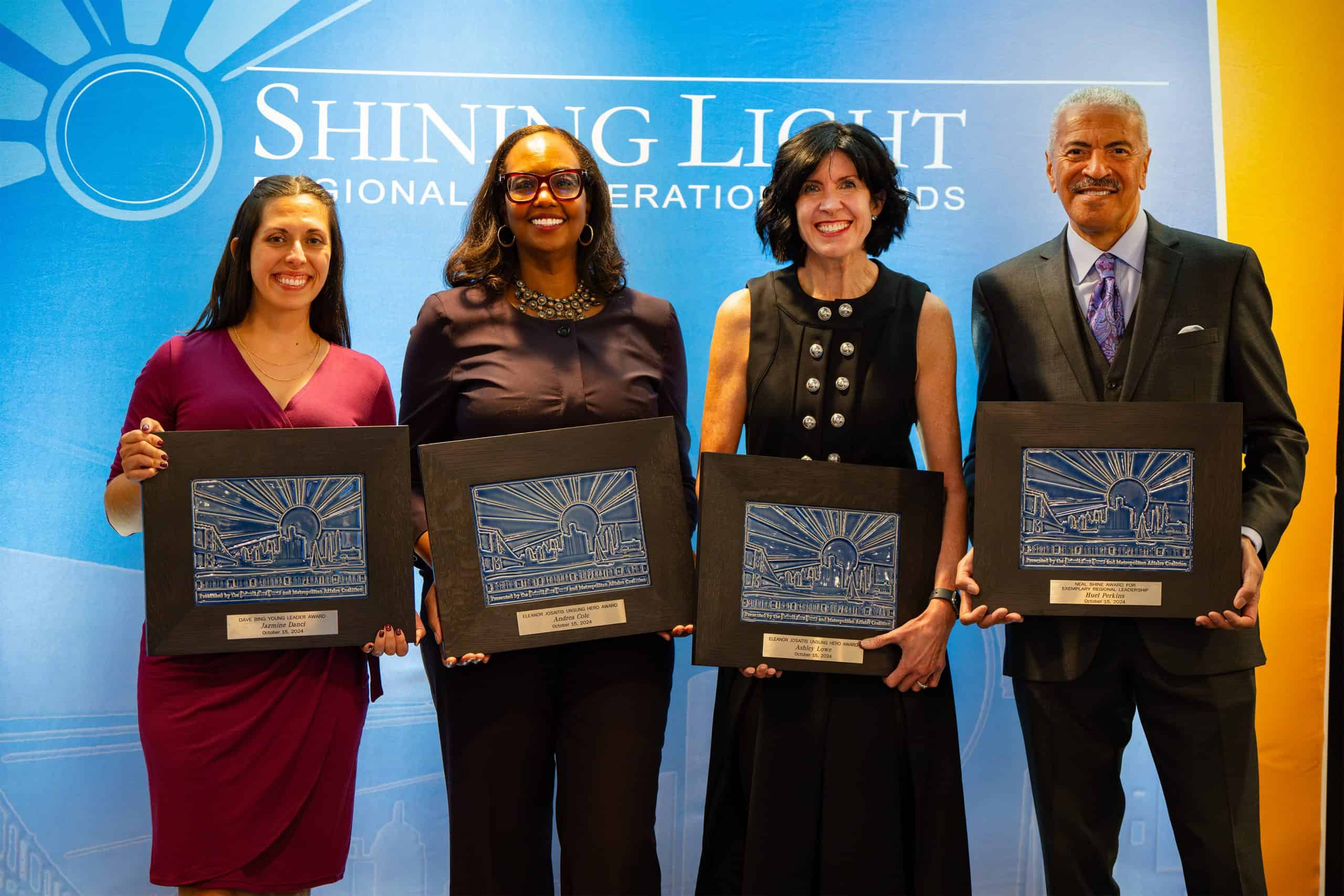 Four award recipients stand in front of a Shining Light Regional Inspiration Awards backdrop, each holding a framed award plaque and smiling at the camera.