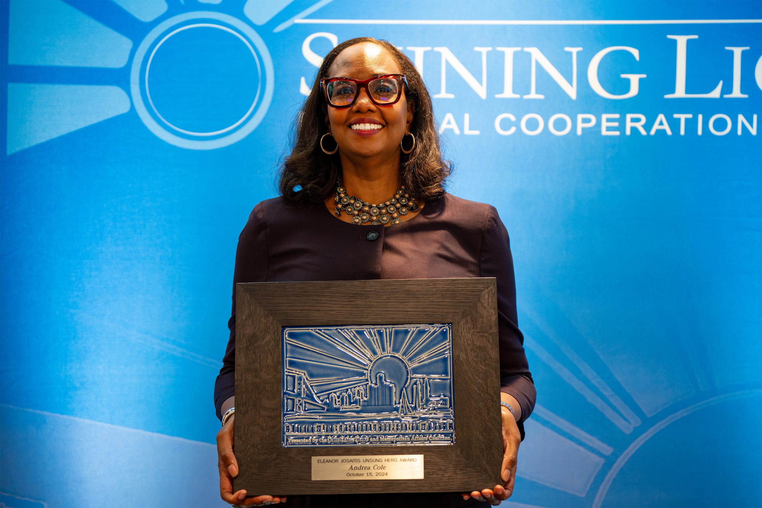 An individual standing in front of a blue backdrop with partially visible text 'Shining Light Regional Cooperation Awards,' holding a framed artwork featuring a cityscape with rays of light. The plaque reads 'Elizabeth Adams Memorial Award Andrea Cole,' indicating an award presentation moment.