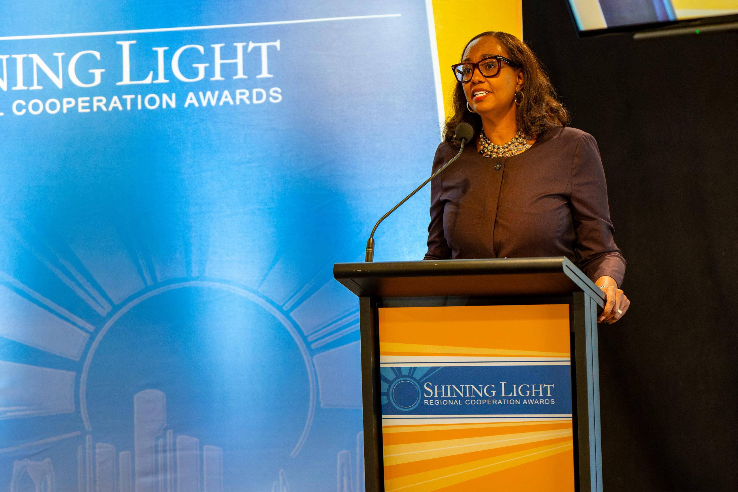 An individual speaking at a podium with a microphone, in front of a backdrop and podium signage that reads 'Shining Light Regional Cooperation Awards,' featuring a sunburst and cityscape illustration. The person is dressed formally, indicating a ceremonial or official event.