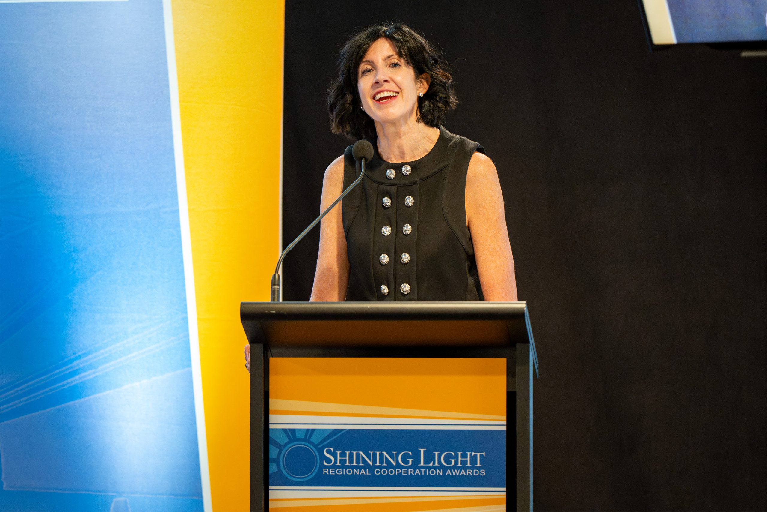 An individual speaking at a podium with a microphone. The podium displays the text 'Shining Light Regional Cooperation Awards,' and the background features blue and yellow colors, indicating a formal event or ceremony.