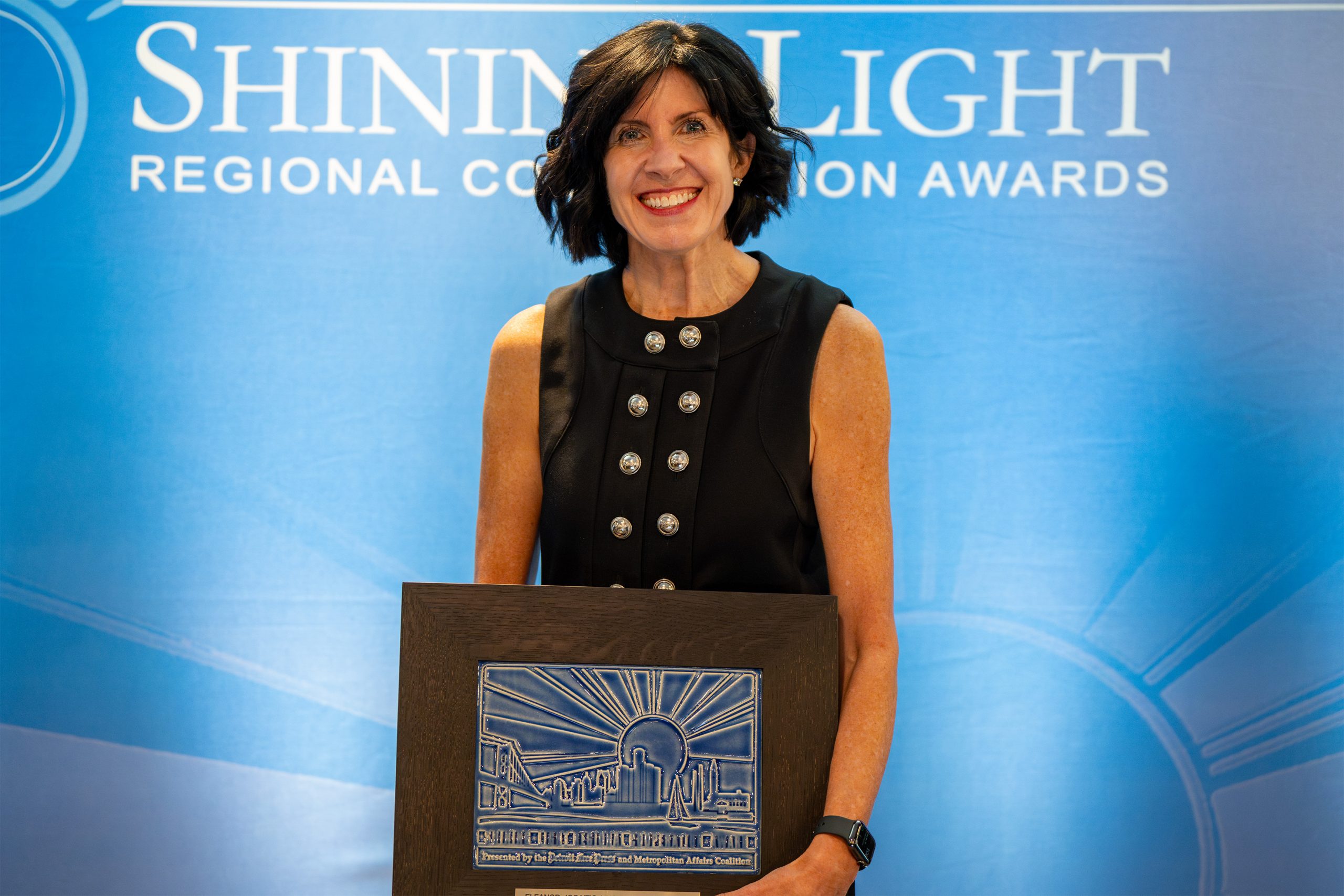 An individual standing in front of a blue backdrop that reads 'Shining Light Regional Cooperation Awards,' holding a plaque with a sunburst and building design. The person is dressed formally, indicating a moment of recognition at an award ceremony.