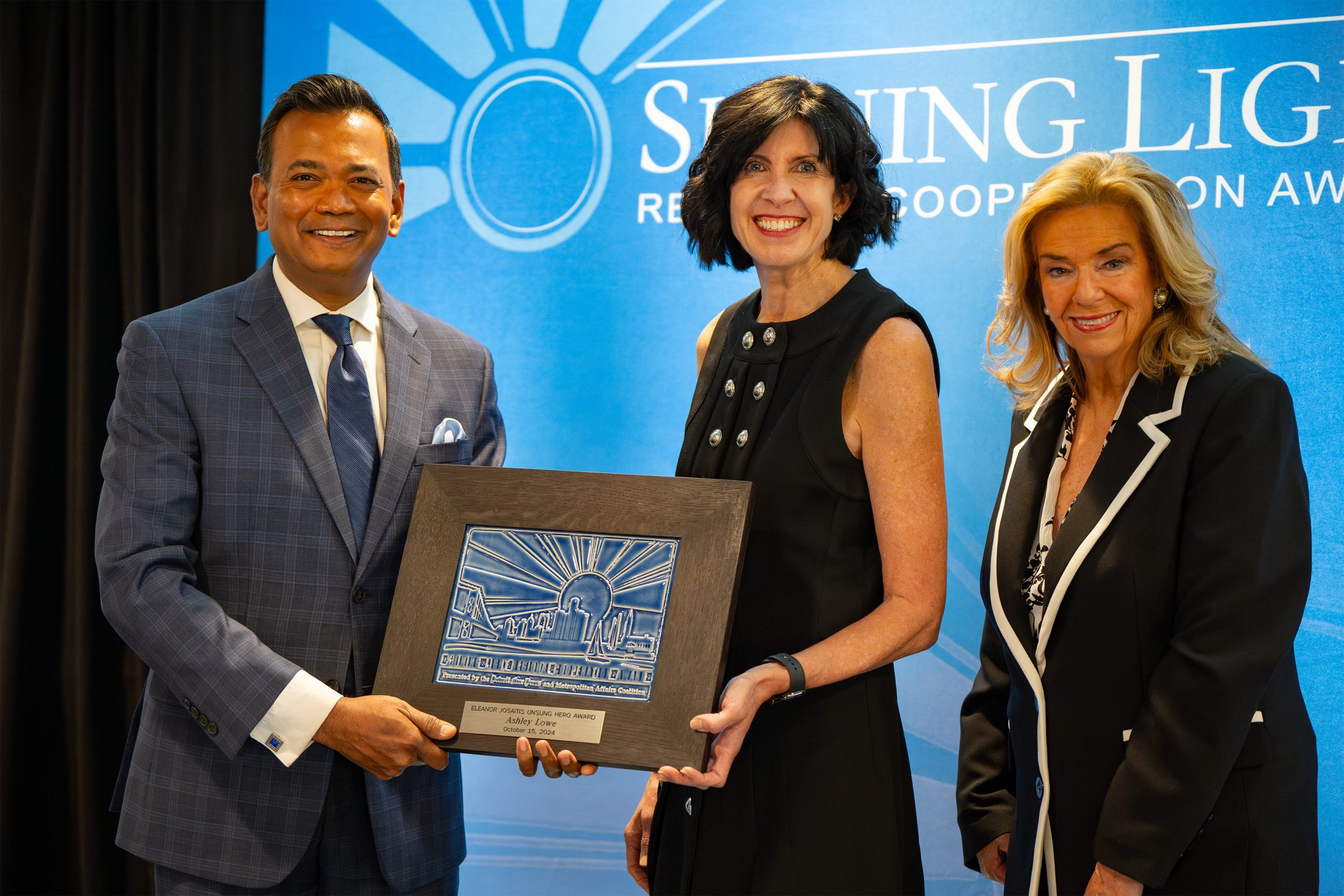 Three individuals standing in front of a blue backdrop with the text 'Shining Light' and 'Regional Cooperation Award.' Two of them are holding a plaque with an engraved design, suggesting an award ceremony recognizing contributions to regional cooperation.