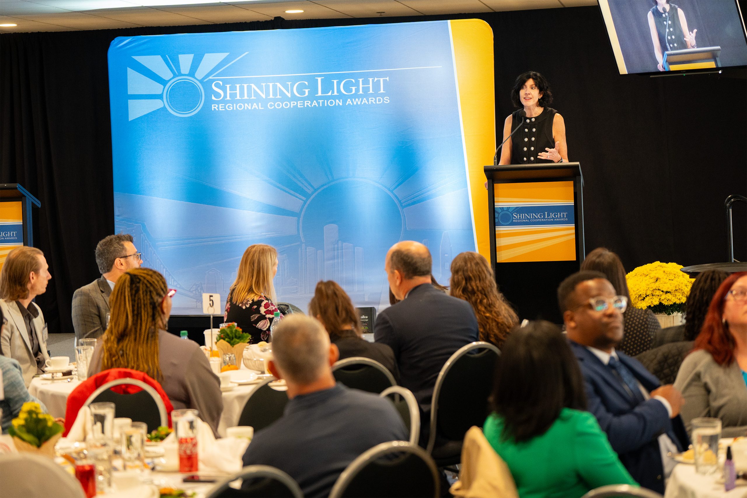 A formal indoor event with attendees seated at round tables, featuring a speaker at a podium on the right. The backdrop reads 'Shining Light Regional Cooperation Awards' with a sunburst and cityscape design. Tables are decorated with yellow flowers, and guests are engaged in conversation or listening.