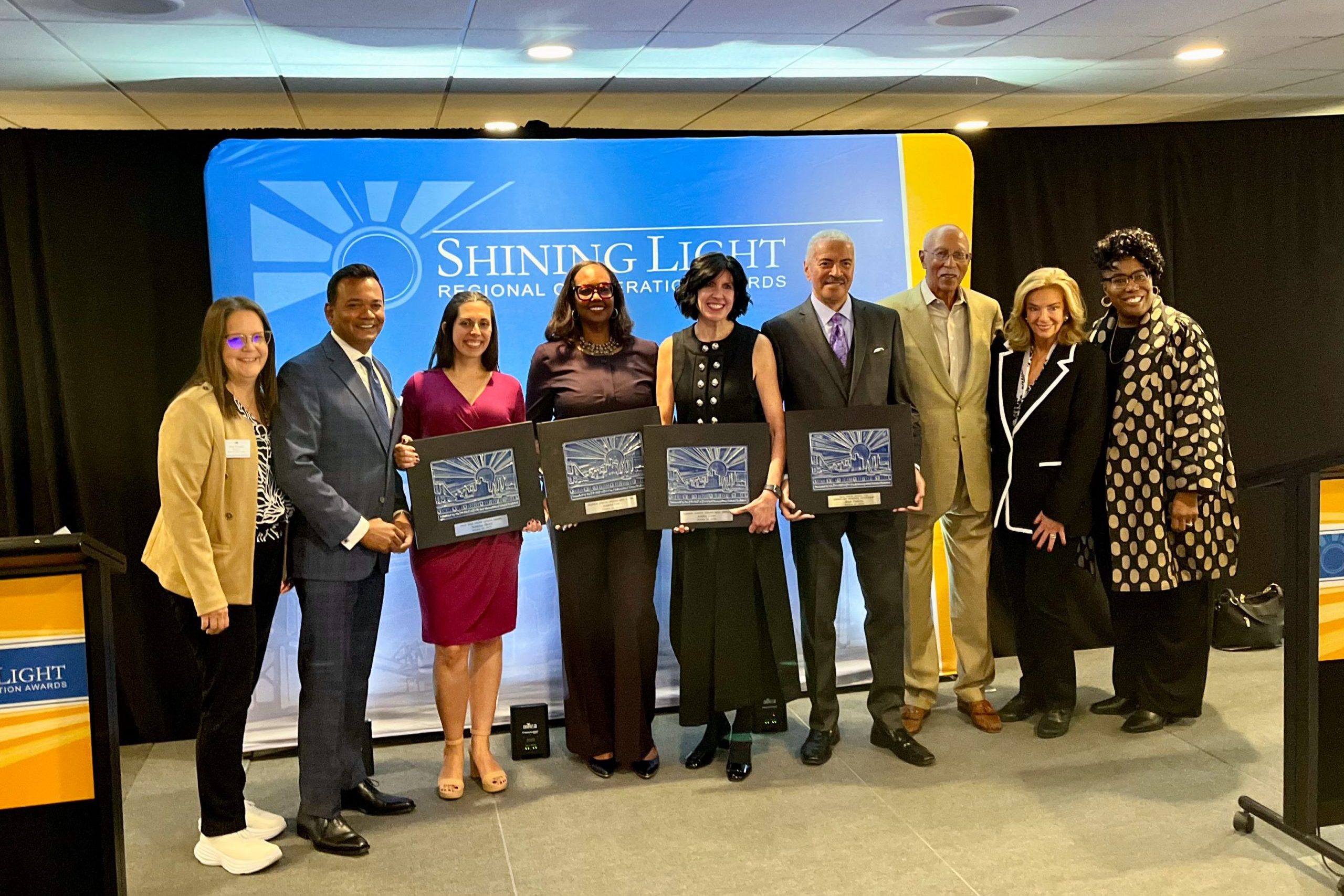 Nine individuals standing in front of a blue and yellow backdrop labeled 'Shining Light Regional Cooperation Awards.' Four people are holding framed awards, indicating a formal recognition ceremony for regional cooperation achievements.