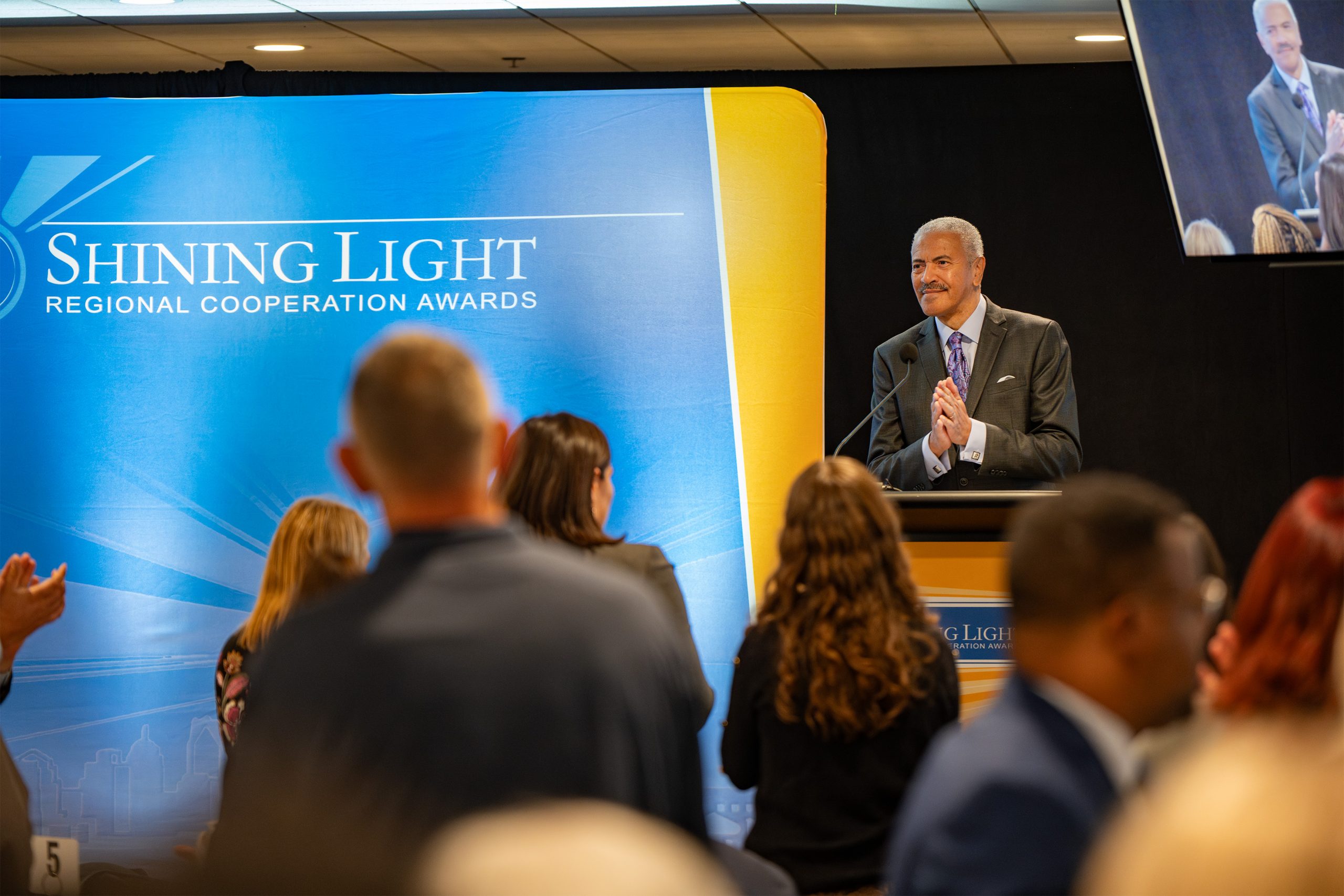 A man standing at a podium clapping, with a large blue and yellow banner behind him reading 'Shining Light Regional Cooperation Awards.' Audience members are visible in the foreground, and a screen in the upper right displays a close-up of the man, indicating a formal award ceremony.
