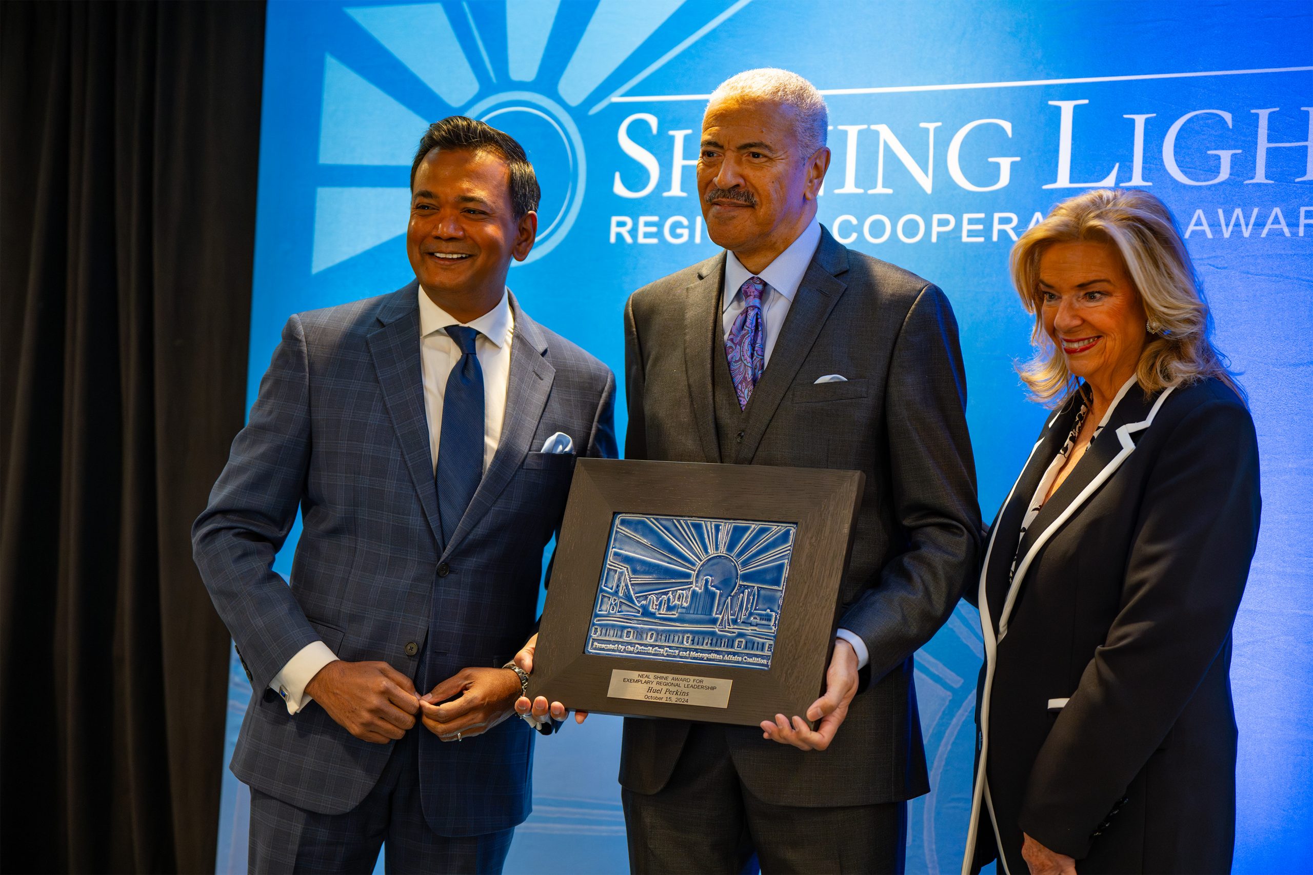 Three individuals standing in front of a blue backdrop that reads 'Shining Light Regional Cooperative Award.' The person in the center holds a plaque with an engraved design featuring rays of light and figures, indicating an award ceremony recognizing regional cooperation.