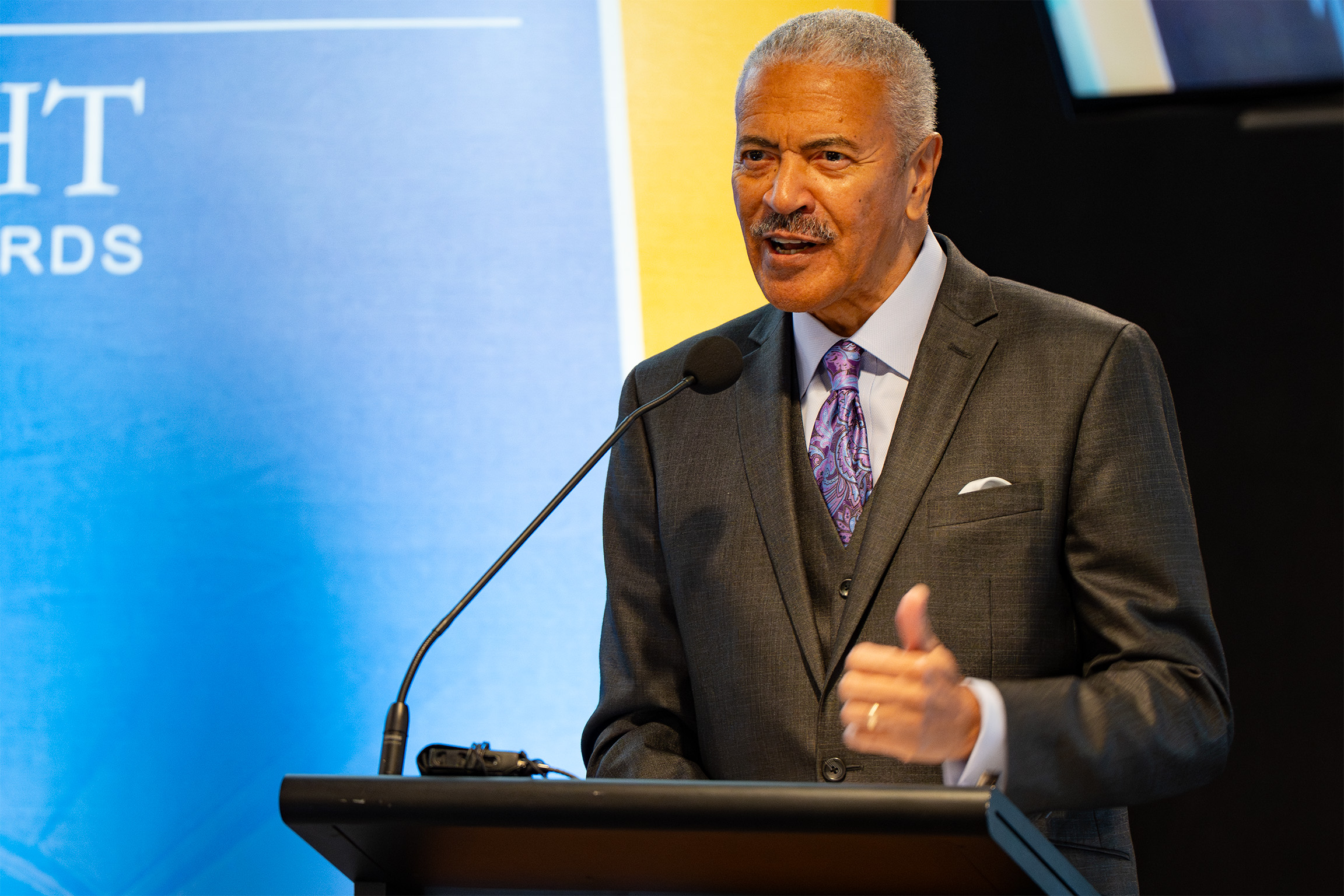 A person in a suit and tie speaking at a podium with a microphone. Behind them is a blue and yellow banner with partially visible text, likely reading 'LIGHT' and 'AWARDS,' suggesting the setting is a formal event or awards ceremony.