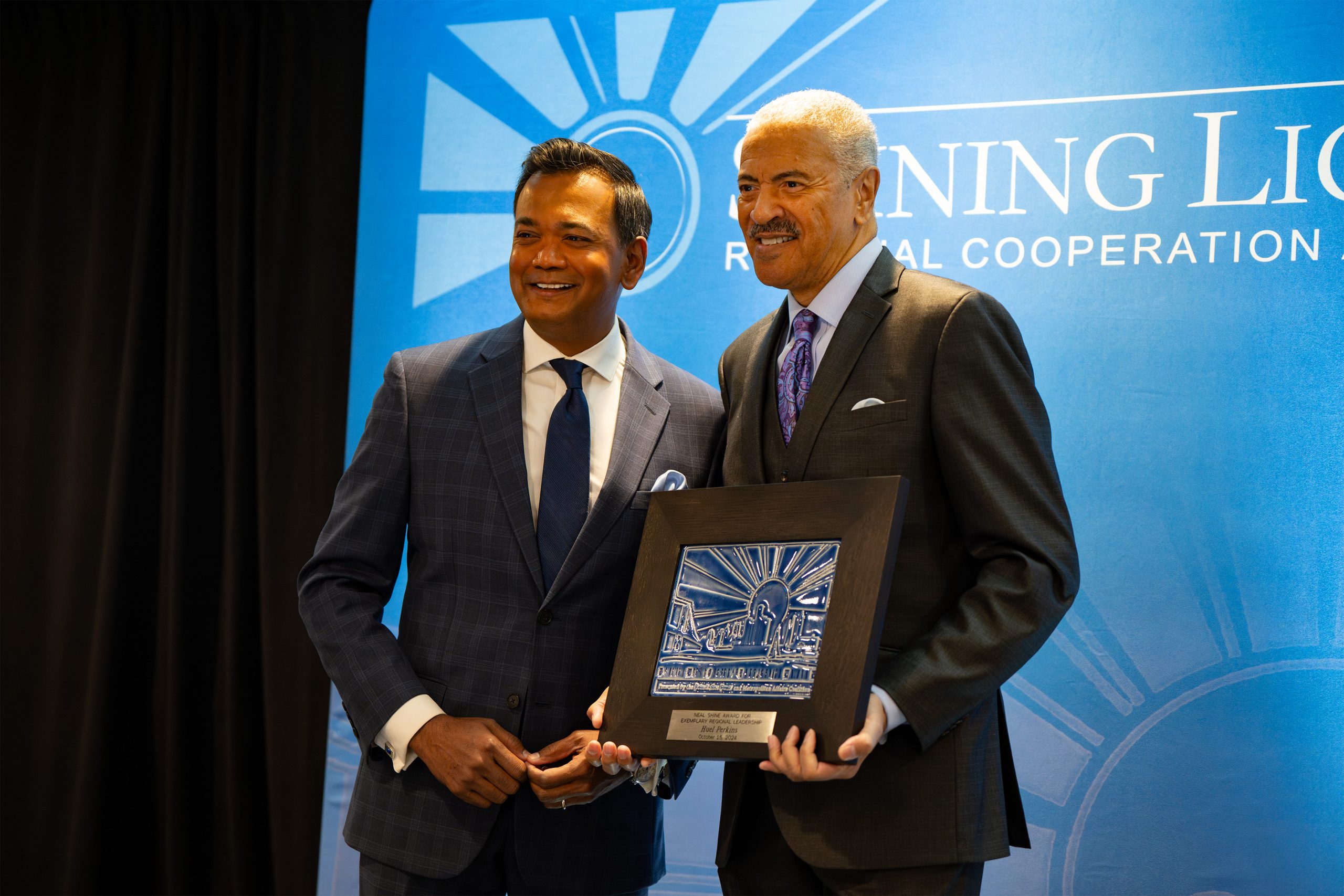 Two individuals standing in front of a blue backdrop with partially visible text 'Shining Light Regional Cooperation.' One person holds a framed award featuring a sunburst design, indicating a recognition moment at a formal event or ceremony.