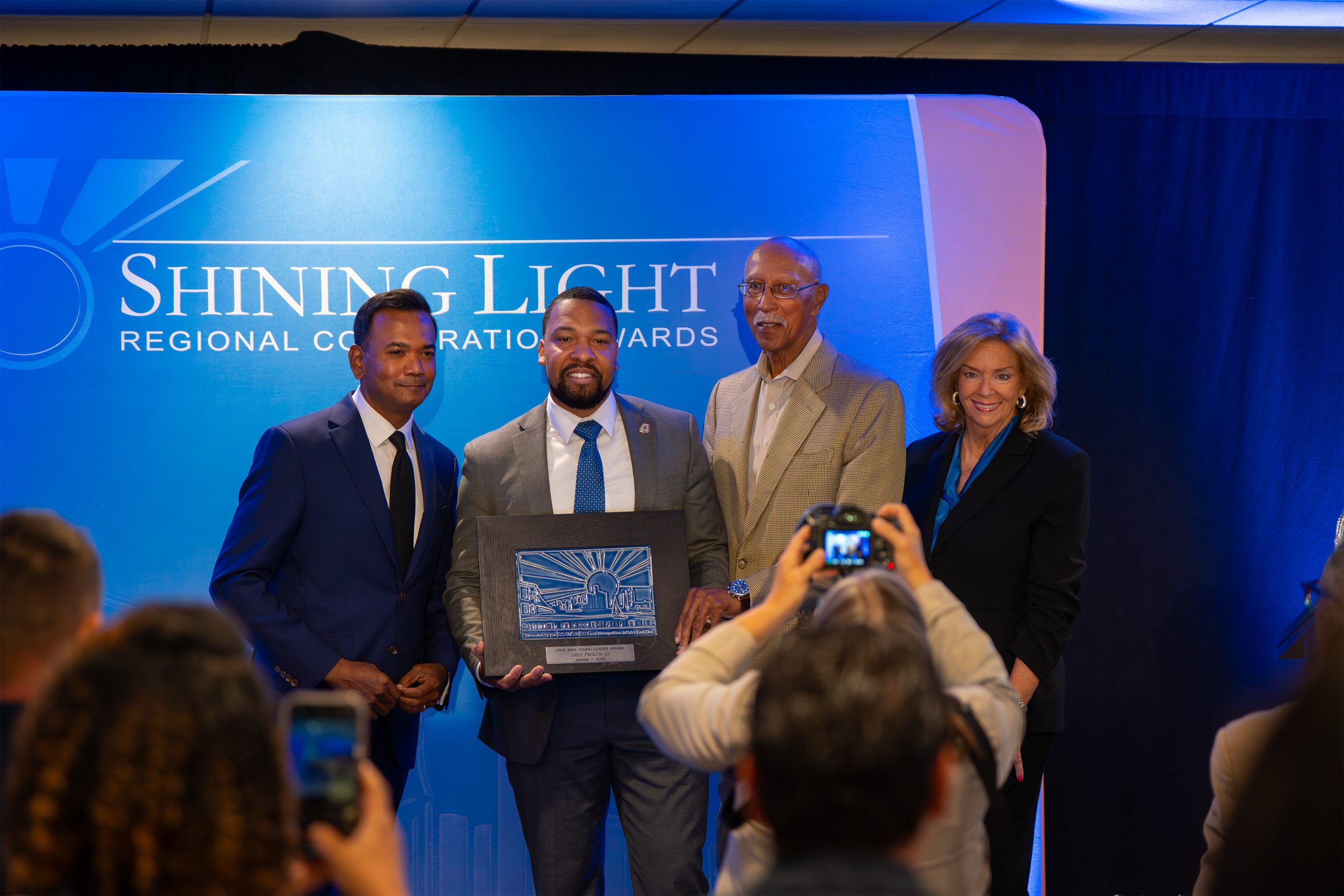 Award ceremony, Shining Light Regional Collaboration Awards, diverse group holding award plaque, professional event highlighting community achievements, recognition, leadership, and teamwork in a formal setting.