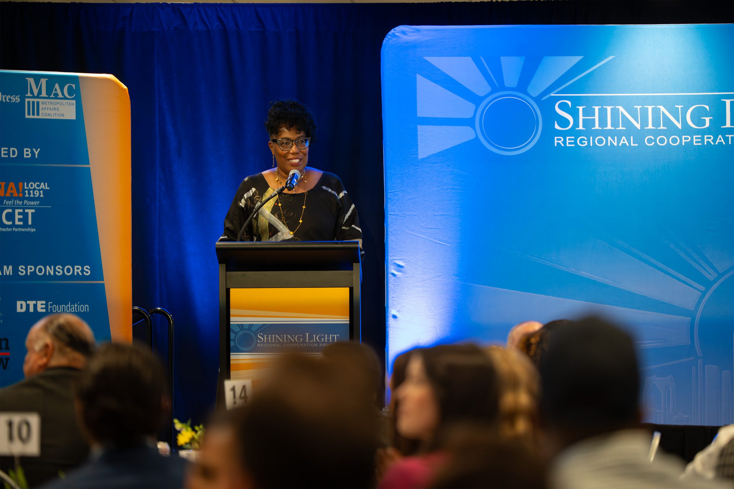 Empowered woman speaking at the Shining Light Regional Cooperation Awards event with blue themed stage and audience in the background.