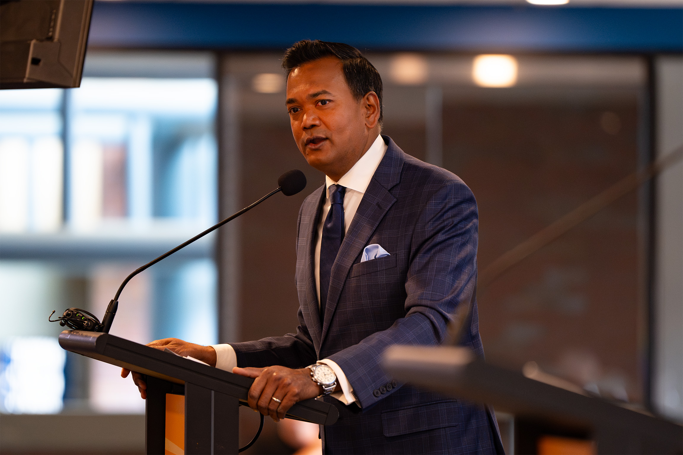 An individual in a suit and tie speaking at a podium with a microphone. The indoor background includes blurred windows and lights, suggesting a formal or professional setting.