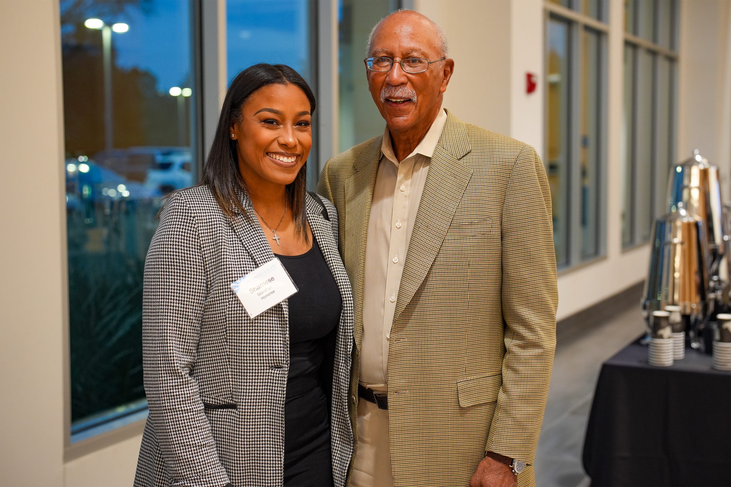 Two individuals in business attire standing indoors at a formal or professional event. One wears a checkered blazer with a name tag, and the other wears glasses and a patterned blazer over a light shirt. Large windows in the background reveal evening lights, and a table with coffee dispensers and cups is visible to the right.