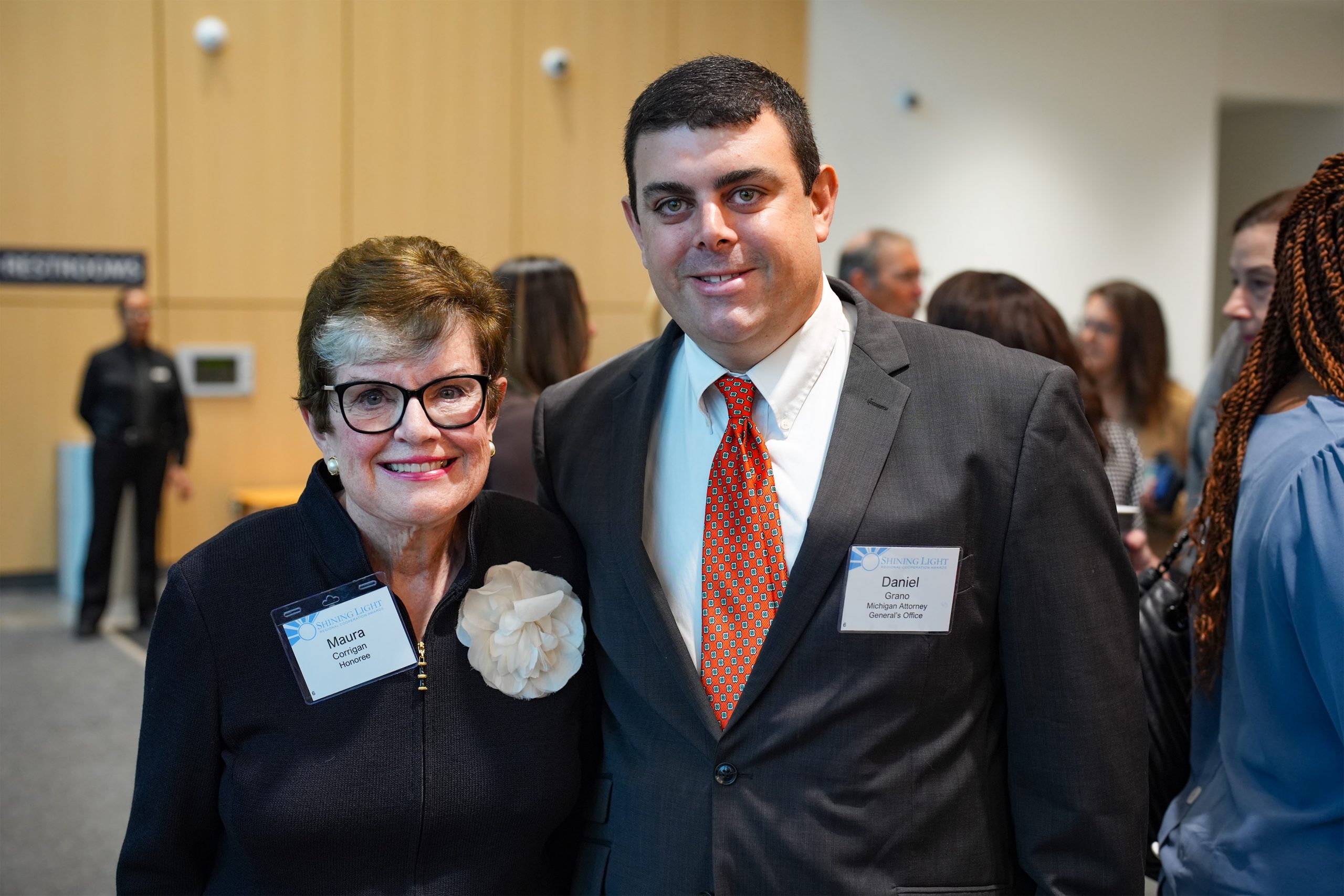 'Two individuals standing side by side at a formal event, both wearing name tags. One wears glasses, a dark outfit, and a large white flower accessory with a name tag reading 'Marian' and 'Trustee.' The other wears a suit with a white shirt and red tie, with a name tag reading 'Darryl.' Other attendees are visible in the background.