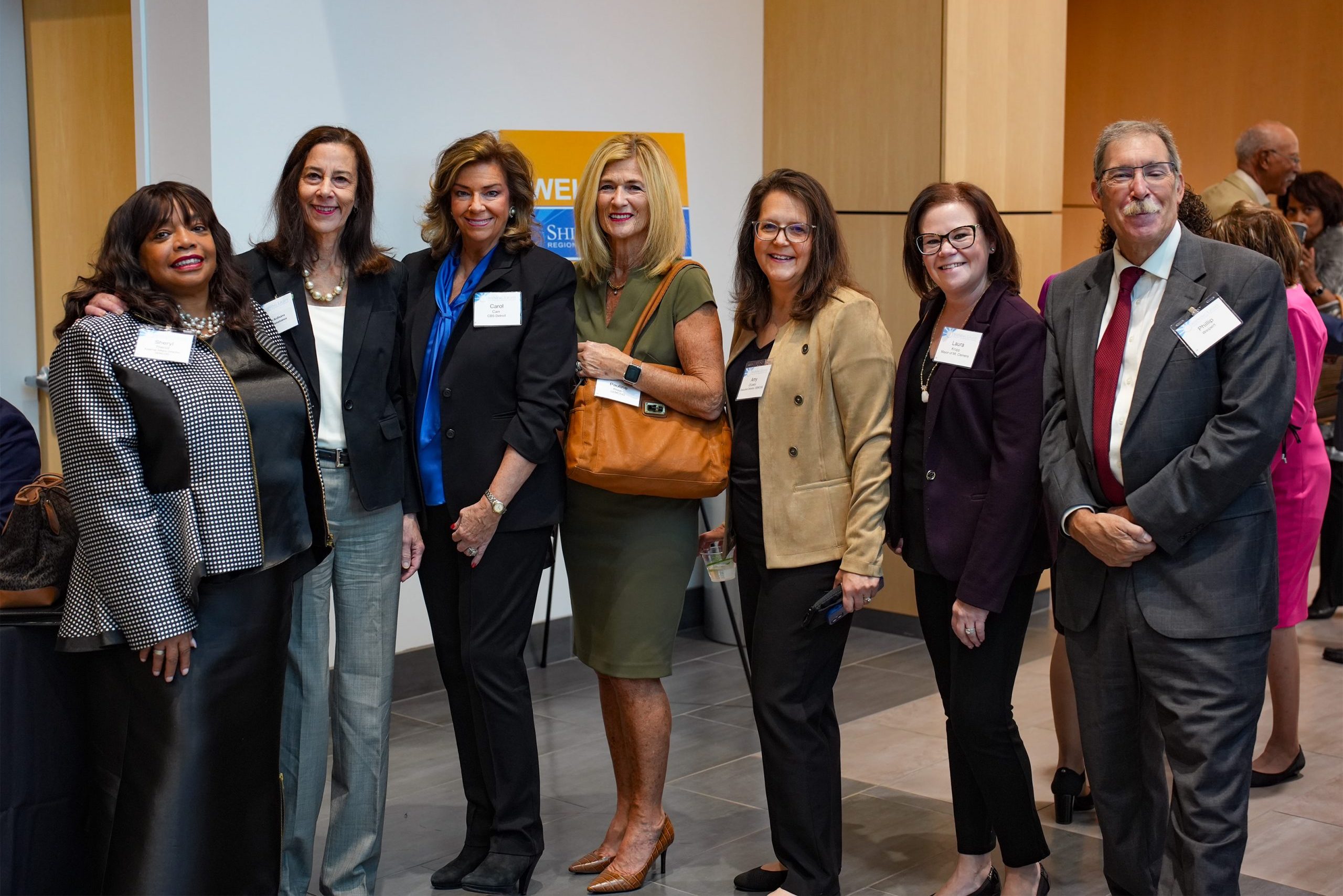 Seven individuals in business attire wearing name tags, standing together in a professional indoor setting.