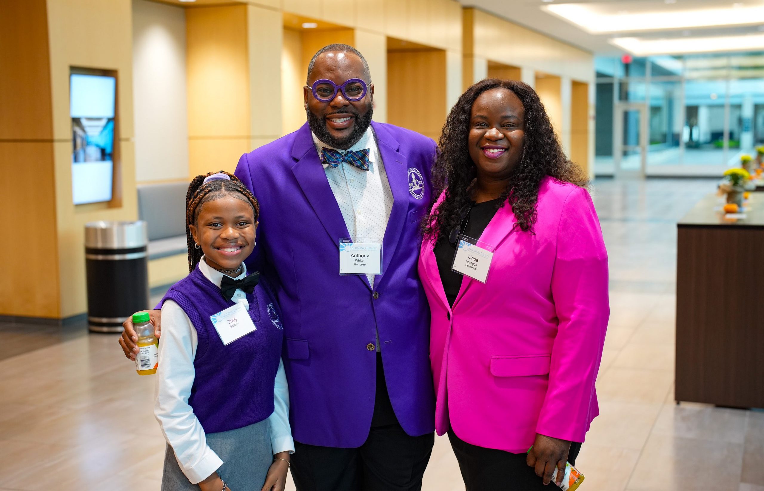 Three individuals standing together indoors, posing for a photo. One wears a white shirt with a purple vest and holds a small bottle; the second wears a purple blazer, white shirt, bow tie, and glasses with a name tag reading 'Andrew'; the third wears a bright pink blazer and a name tag. All are smiling.
