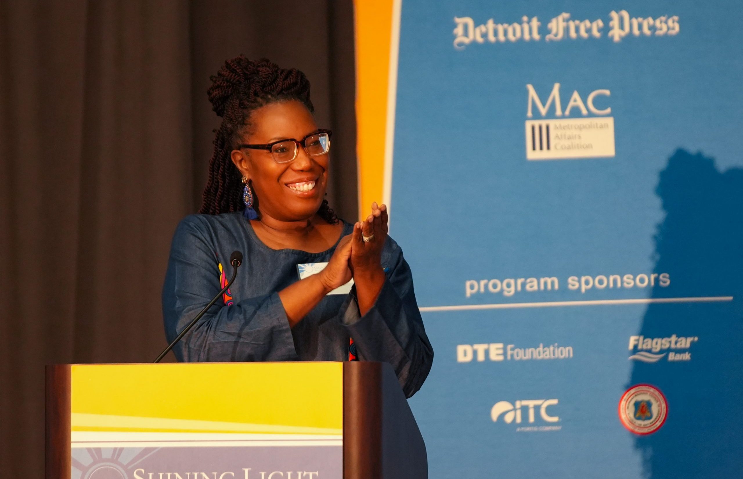 A person standing at a podium labeled 'Shining Light Regional Cooperation Awards,' clapping and smiling. Behind them is a blue banner displaying 'Detroit Free Press,' 'MAC Metropolitan Affairs Coalition,' and sponsor logos including DTE Foundation, Flagstar Bank, and ITC Holdings Corp.