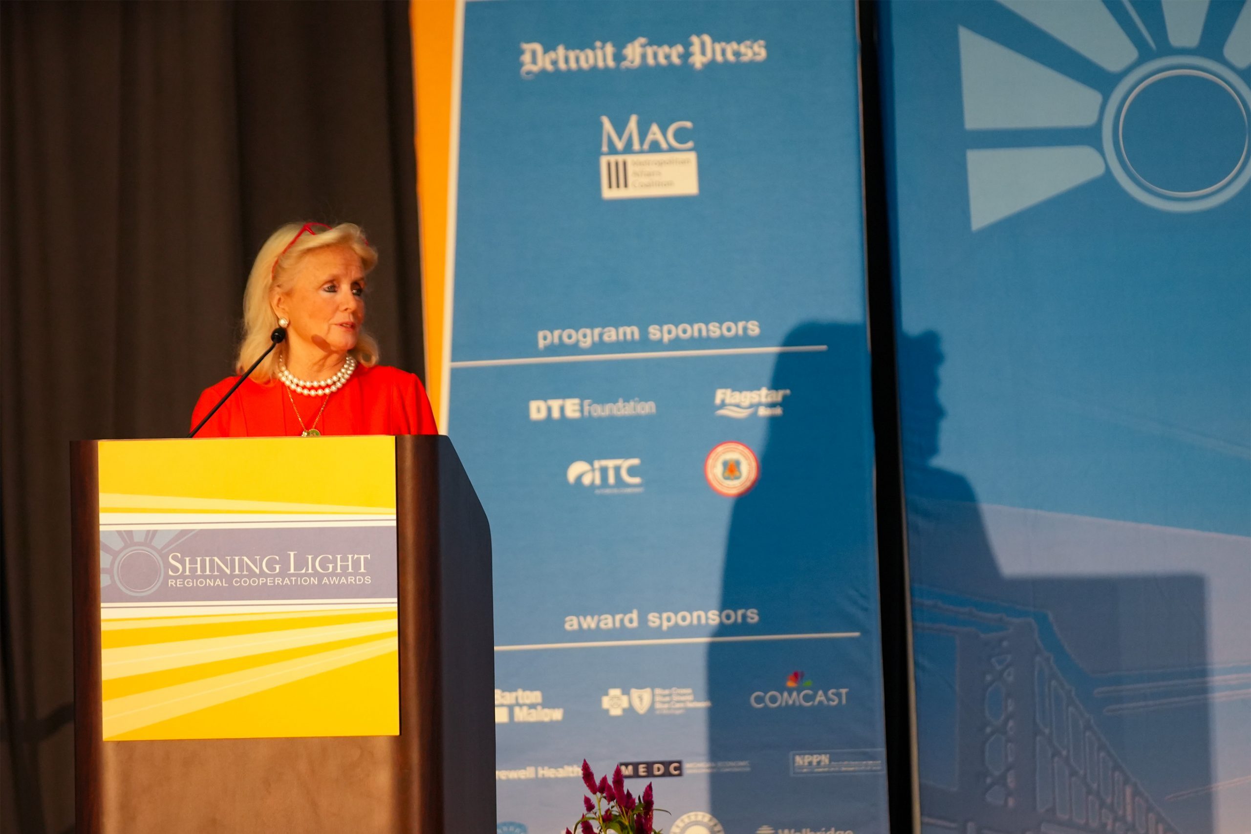 A person standing at a podium labeled 'Shining Light Regional Cooperation Awards,' with a blue backdrop displaying sponsor logos including Detroit Free Press, MAC, DTE Foundation, Flagstar Bank, ITC Holdings Corp., Barton Malow, Blue Cross Blue Shield of Michigan, Comcast, Henry Ford Health System, MEDC, and Walbridge. Flowers are arranged in front of the podium.