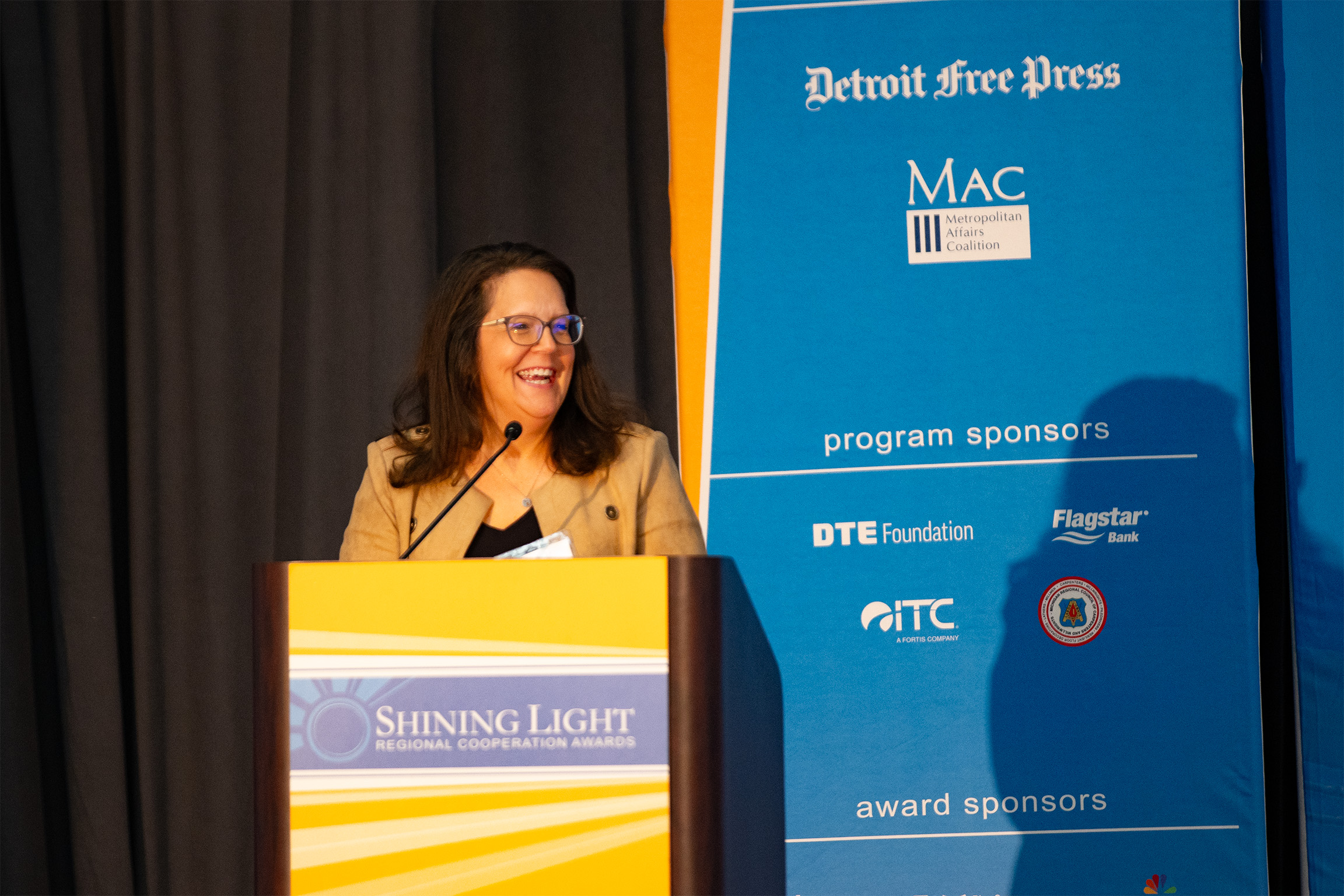 A person speaking at a podium during the Shining Light Regional Cooperation Awards. The podium features the event name, and the backdrop behind them displays sponsor logos and text including Detroit Free Press, MAC Metropolitan Affairs Coalition, DTE Foundation, and Flagstar Bank.