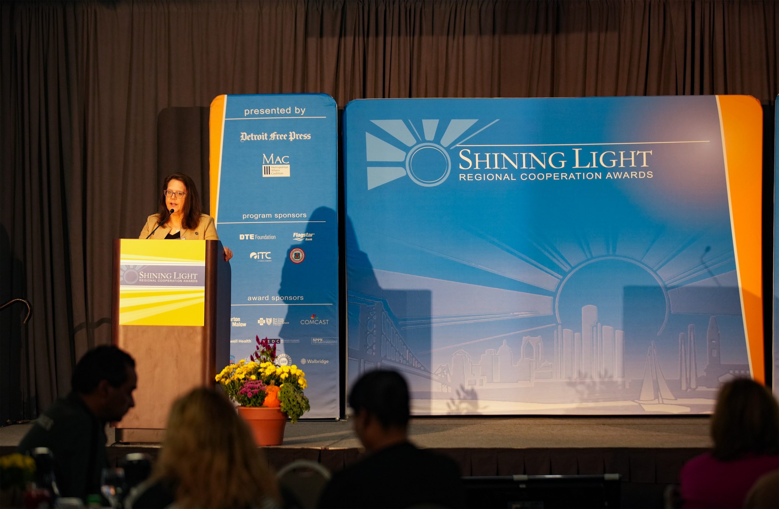 'Stage setup for the Shining Light Regional Cooperation Awards with a speaker at a podium on the left side. The podium and backdrop display the event's name and logo. A vertical banner lists sponsors including Detroit Free Press, MAC, DTE Energy Foundation, and ITC Holdings Corp. Potted flowers decorate the front of the podium, and attendees are seated facing the stage.