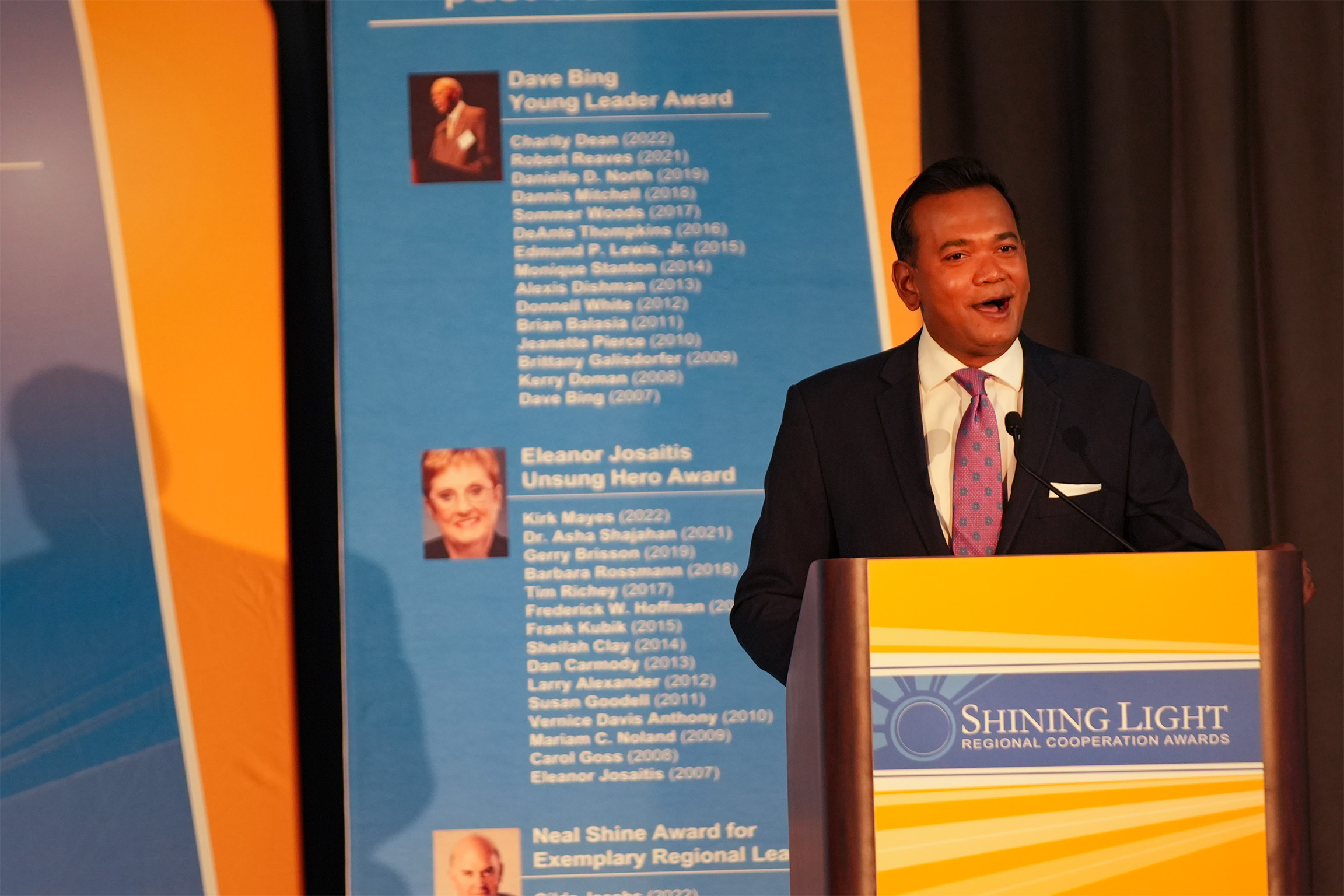 Man in suit speaking at a podium with a blue backdrop reading ‘Shining Light Regional Cooperation Awards.’ Behind him is a tall sign listing award names such as ‘Dave Bing Young Leader Award’ and ‘Eleanor Josaitis Unsung Hero Award.’