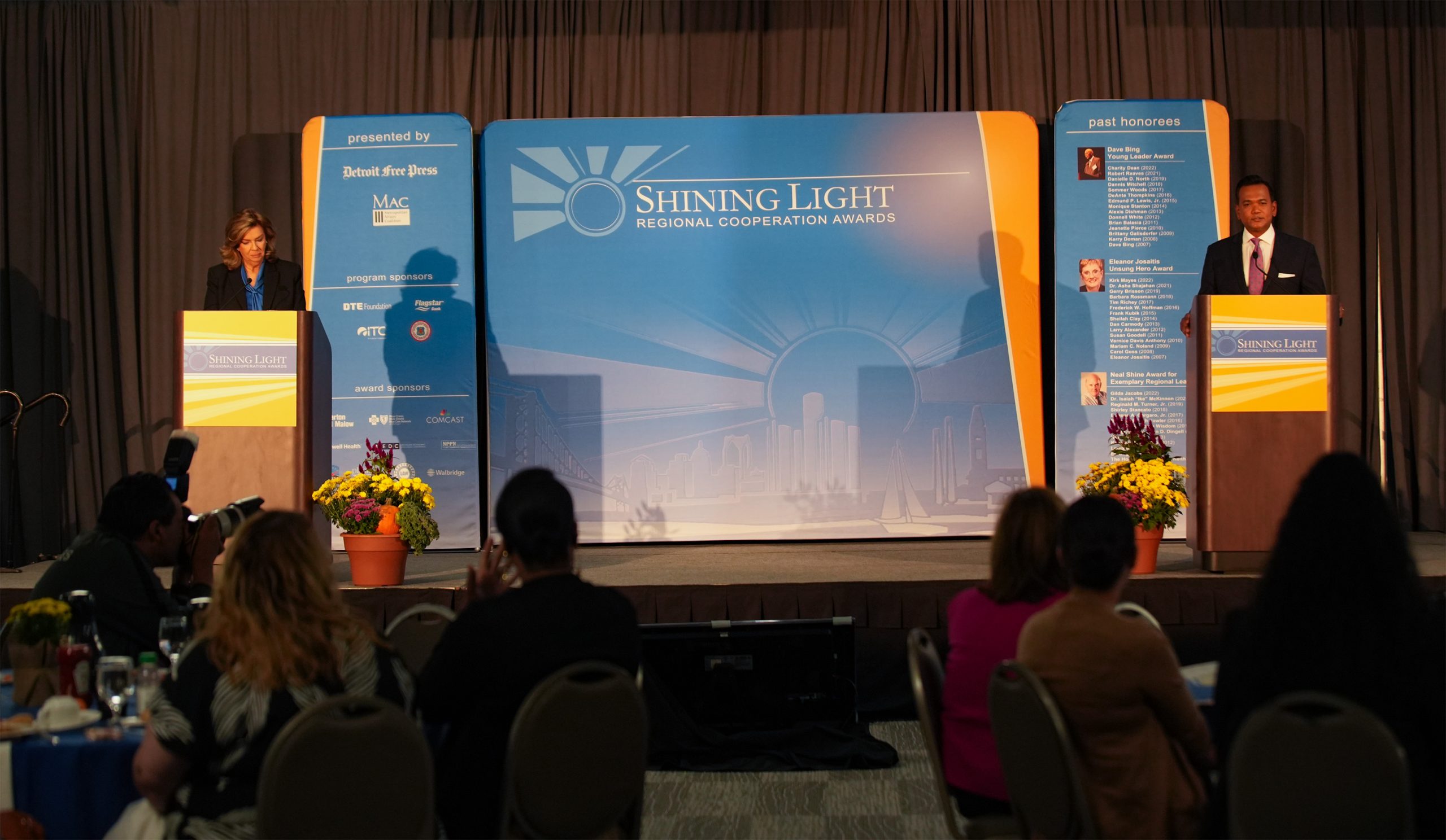 Stage setup for the Shining Light Regional Cooperation Awards with two podiums, each occupied by a speaker. The central backdrop displays the event name and logo, flanked by tall banners listing sponsors and past honorees. Audience members are seated at round tables in front of the stage in a formal setting.