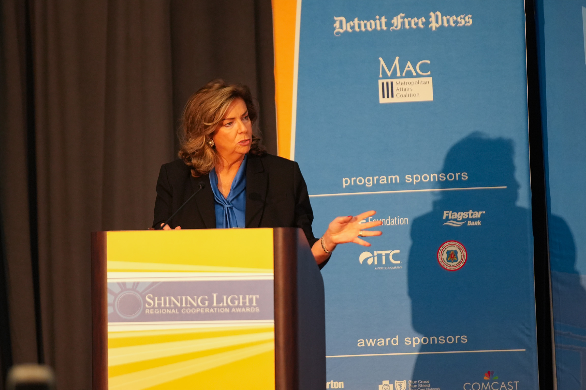 A person standing at a podium giving a speech at the Shining Light Regional Cooperation Awards. The podium sign displays the event name, and a blue backdrop behind them features logos and sponsor names including Detroit Free Press, MAC Metropolitan Affairs Coalition, Flagstar Bank, and ITC.