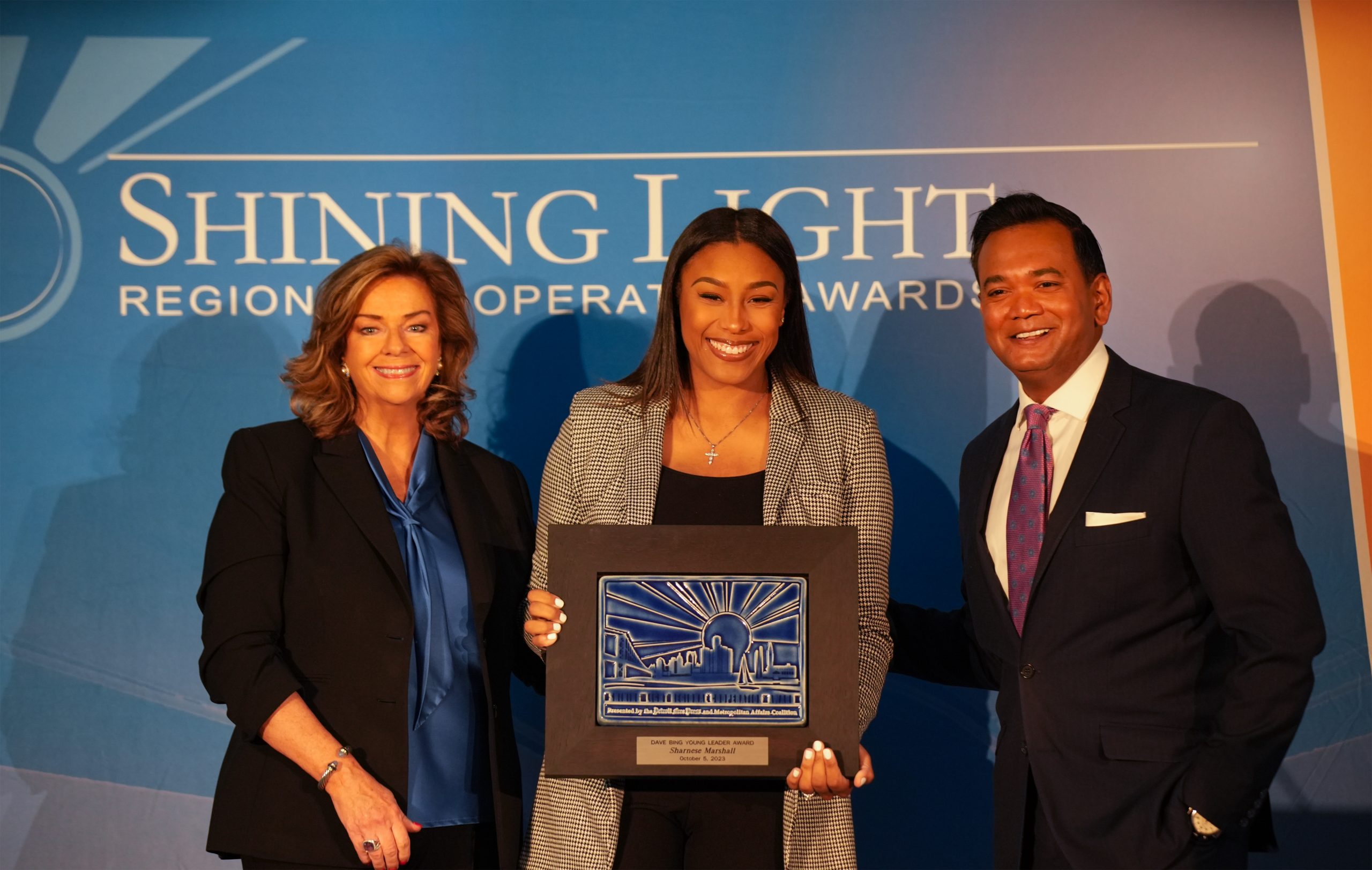Three people standing side-by-side on a stage holding plaques in front of a blue backdrop that reads ‘Shining Light Regional Cooperation Awards.