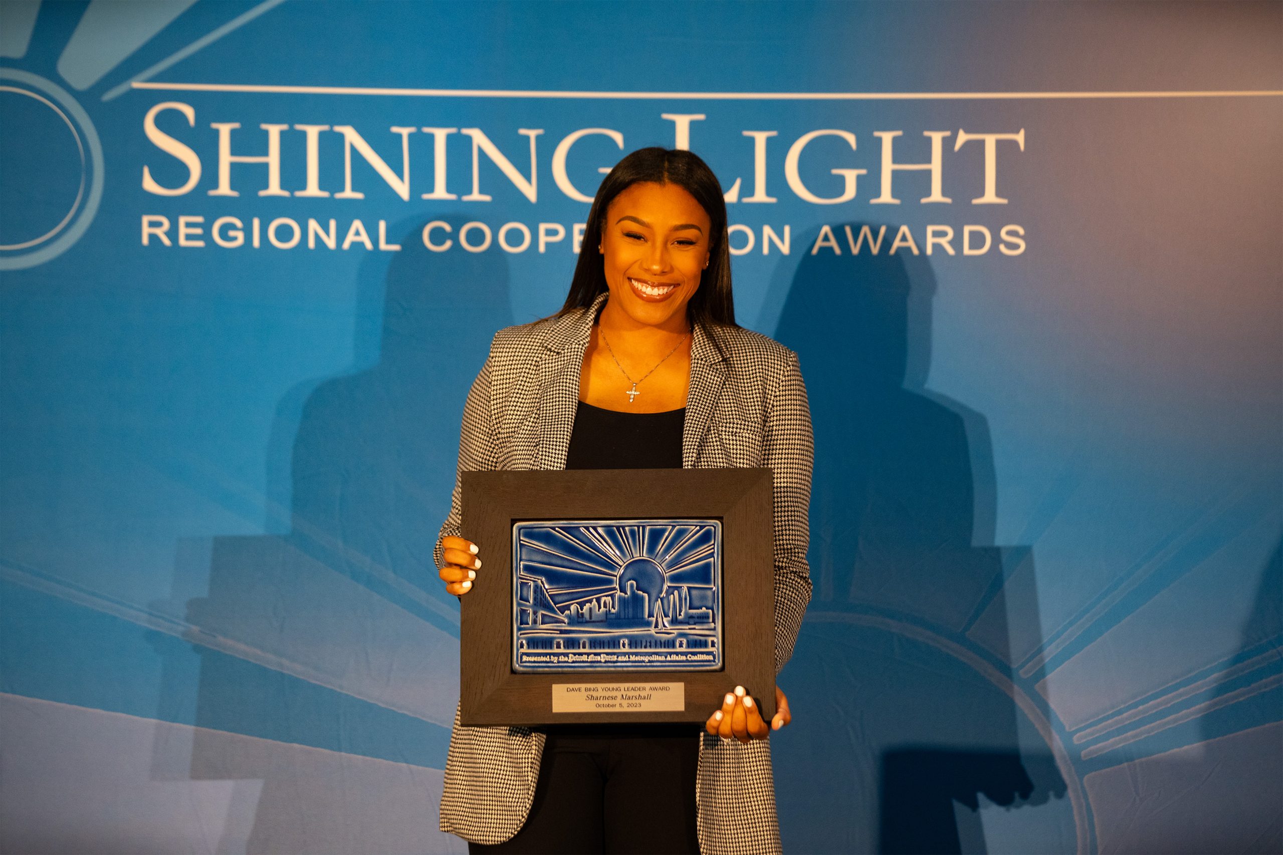 A person standing in front of a blue backdrop labeled 'Shining Light Regional Cooperation Awards,' holding a framed award featuring a cityscape design with rays and a plaque that reads 'For Outstanding Contribution to the Community.