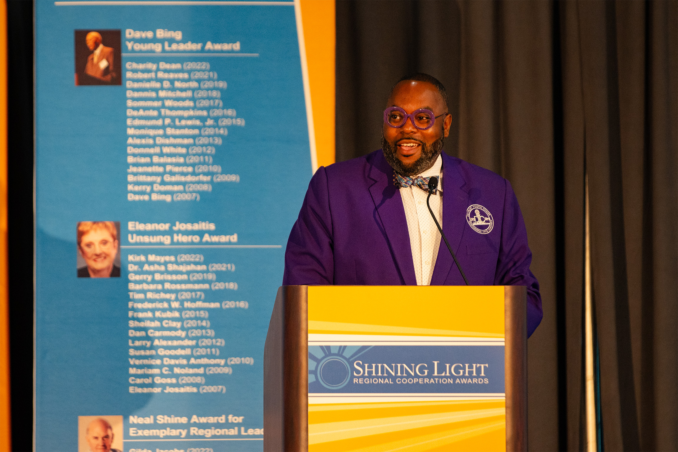 A person wearing glasses and a purple jacket with an emblem speaks at a podium labeled 'Shining Light Regional Cooperation Awards.' Behind them is a blue and yellow banner listing past recipients of the Dave Bing Young Leader Award, Eleanor Josaitis Unsung Hero Award, and Neal Shine Award for Exemplary Regional Leadership.