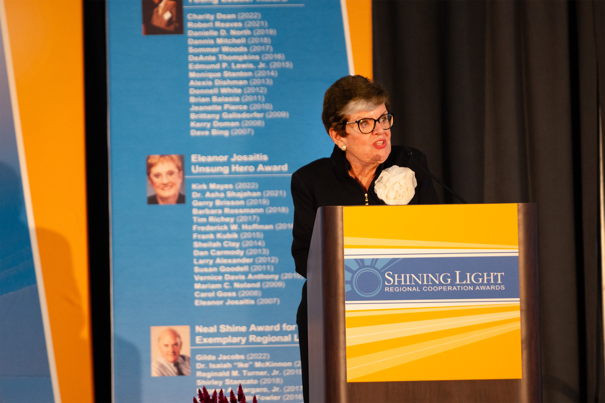 A person speaking at a podium during the Shining Light Regional Cooperation Awards. Behind them is a blue and yellow banner listing past award recipients under categories like the Eleanor Josaitis Unsung Hero Award and Neal Shine Award for Exemplary Regional Leadership, with names and years including Charity Dean (2022), Kirk Mayes (2022), Dr. Asha Shajahan (2021), and Gilda Jacobs (2022).