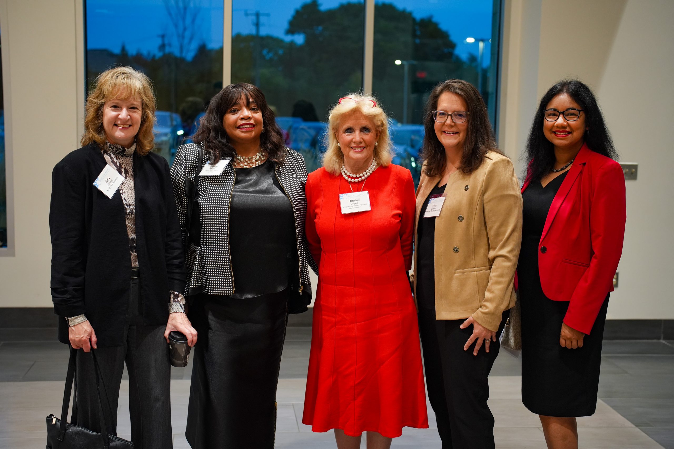 Five individuals in formal attire posing for a group photo indoors, all wearing name tags. Large windows in the background reveal trees and buildings, suggesting the photo was taken during an event or conference