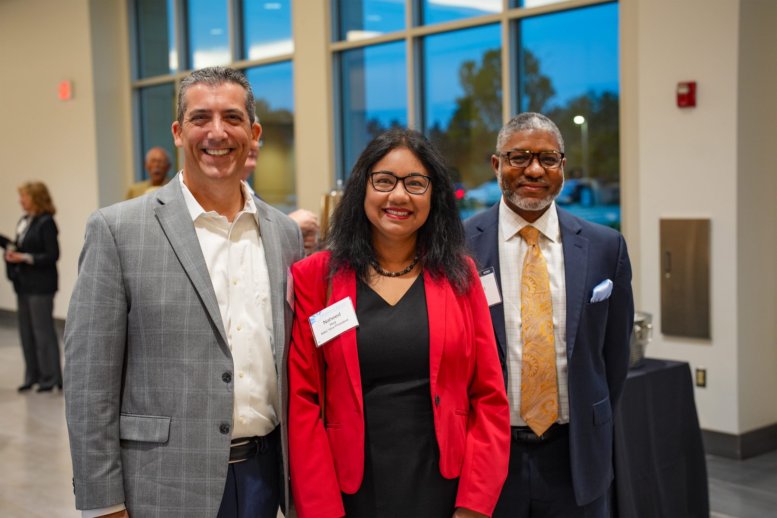 Three individuals in business attire standing together indoors in front of large windows, possibly during a professional event. Two wear suits and one wears a red blazer. Two of them have visible name tags, suggesting a conference or networking setting.