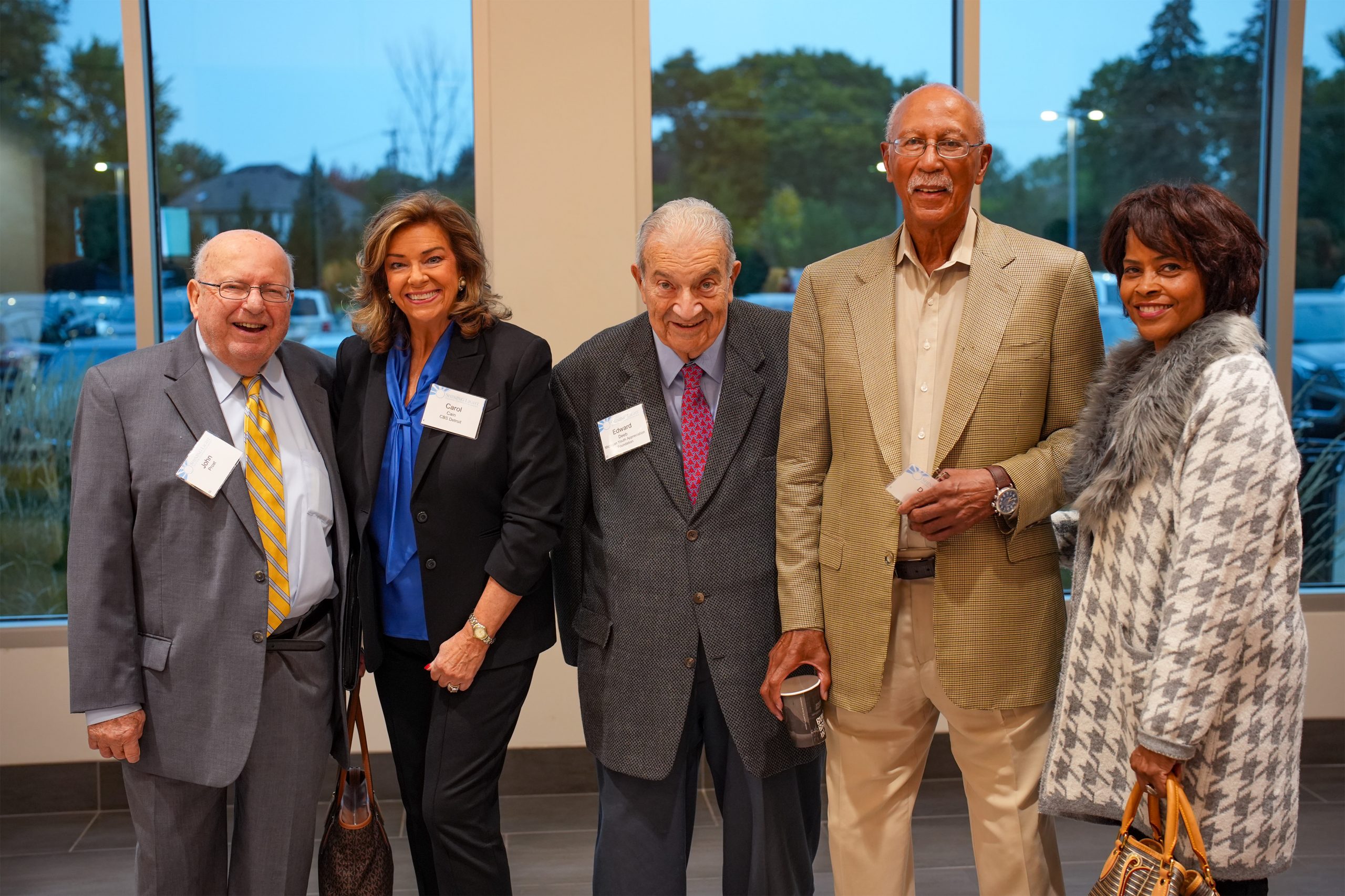 Five individuals standing indoors in front of large windows, dressed in formal and semi-formal attire with name tags. The group includes three people in suits and ties, one in a blue shirt and black jacket, and one in a patterned coat with fur trim. A parking lot and trees are visible through the windows.