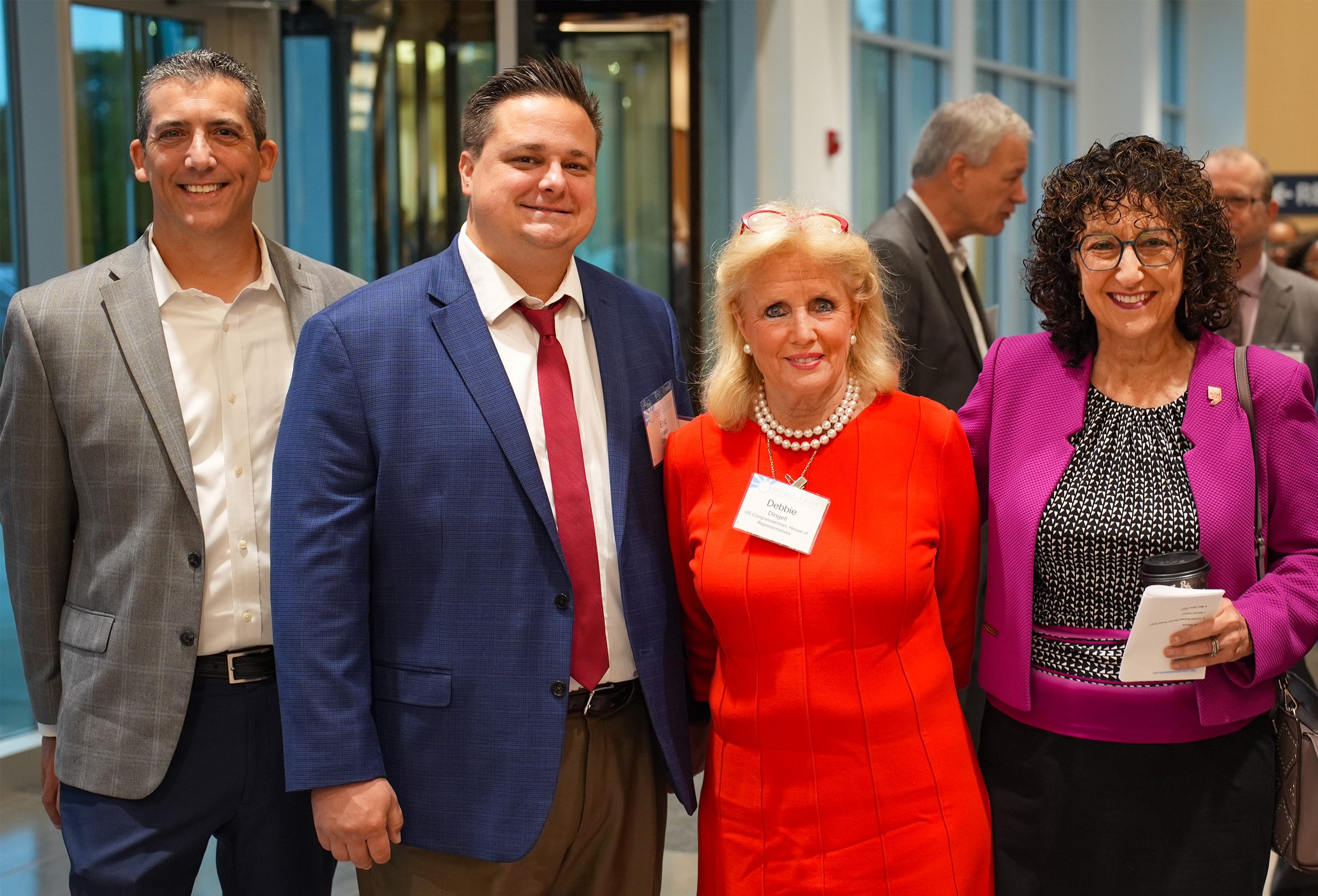 Four individuals in formal attire standing together indoors at a professional or social event. One wears a gray suit, another a blue suit with a red tie, the third wears a red dress with pearls and a name tag reading 'Debbie,' and the fourth wears glasses and a purple jacket over black attire, holding papers. Large windows and other attendees are visible in the background.
