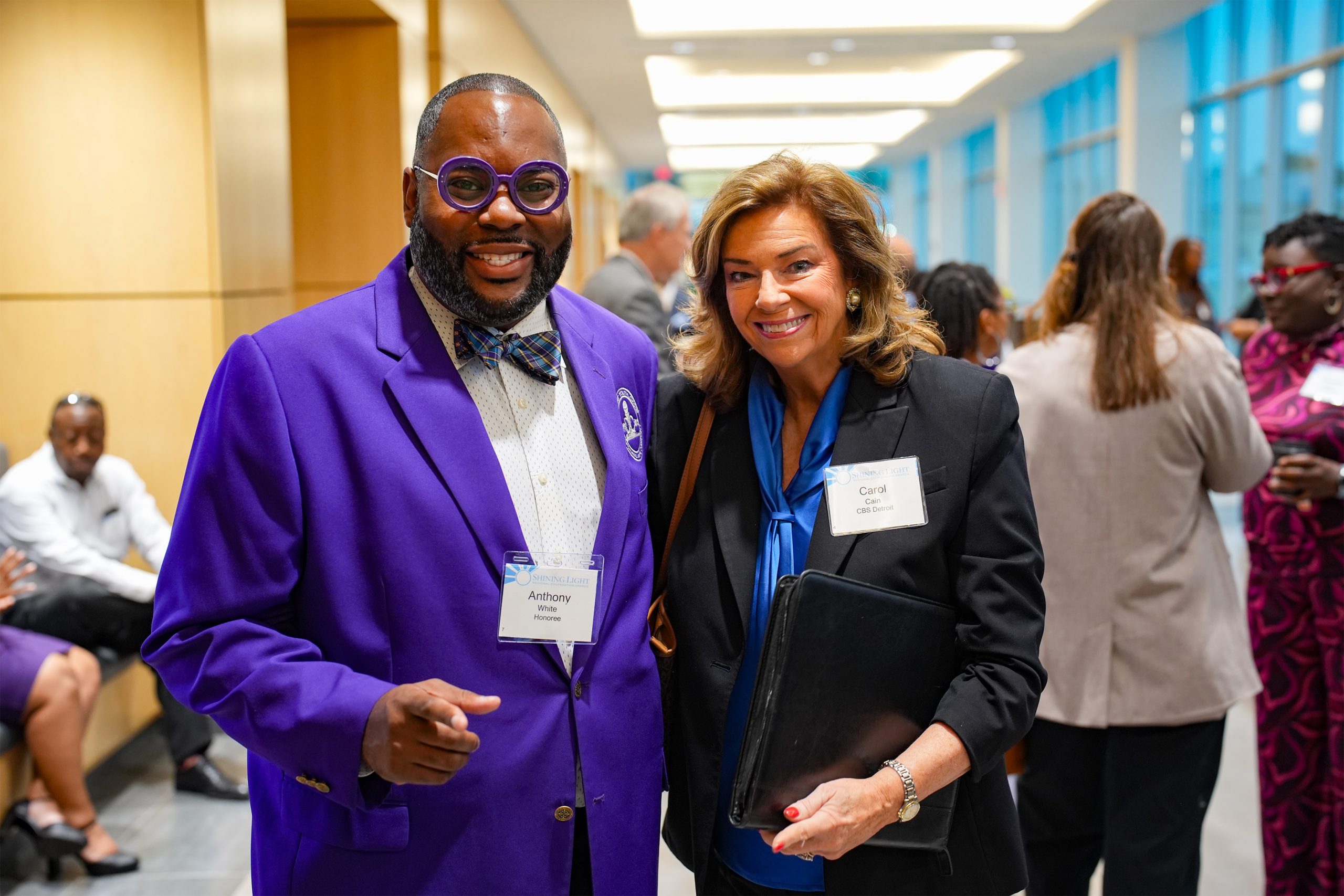 Two individuals standing side by side in a brightly lit indoor setting, possibly a professional event. One wears a purple suit with a bow tie and glasses, and the other is in business attire holding a folder. Both have name tags. Other attendees are visible in the background, engaged in conversation.