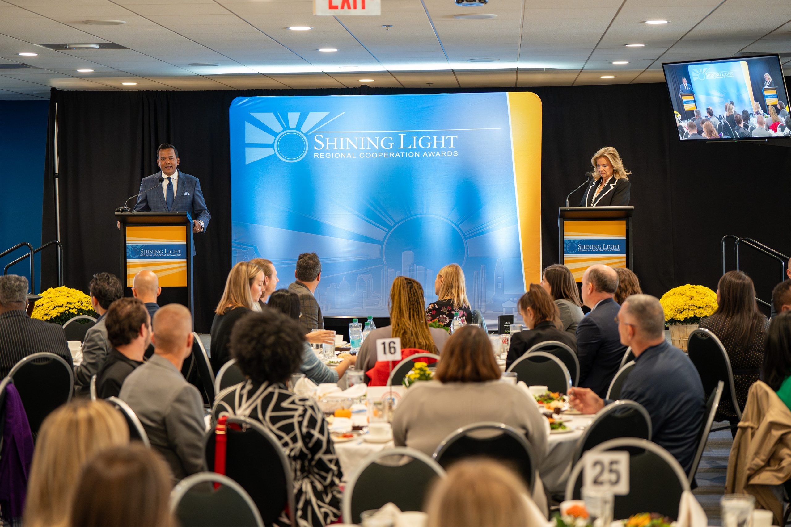 Audience seated at round tables during the Shining Light Regional Cooperation Awards event, watching two speakers at podiums on stage. The backdrop features a sunburst and cityscape graphic with the event name. Table numbers like '16' and '25' are visible, and a screen displays the scene from another angle.