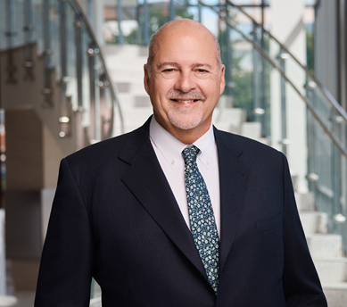 Professional portrait of an individual wearing a dark business suit, white dress shirt, and patterned tie, standing in a modern office building interior. A staircase with glass railings rises in the background, and large windows provide natural light throughout the space. The image highlights leadership, professionalism, and a corporate or organizational setting.