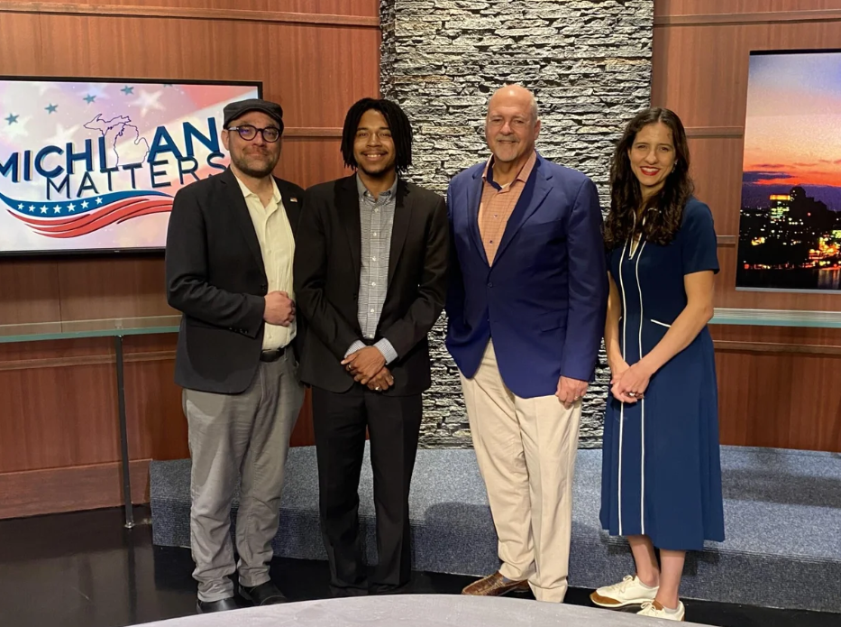 Four individuals stand together on a television studio set in front of Michigan Matters program display screens.